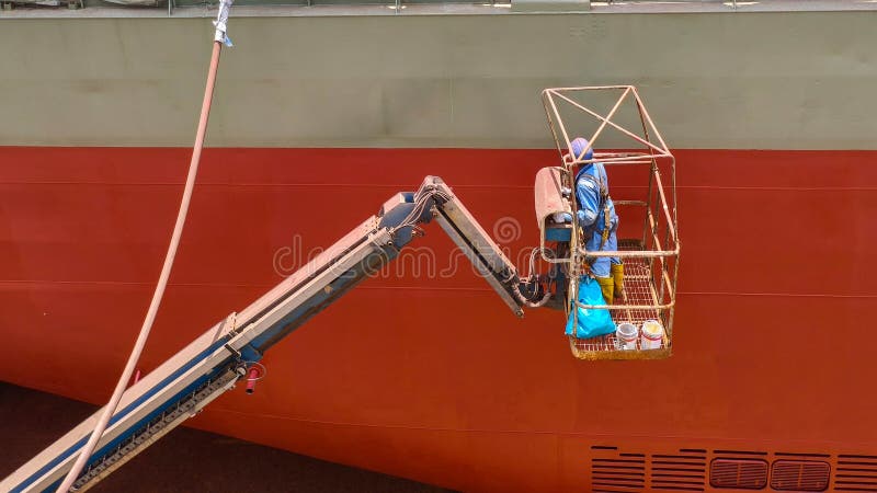 Large ship in a dry dock undergoing maintenance. A worker is in an elevated platform attached to a crane, attending to the ship. stock image