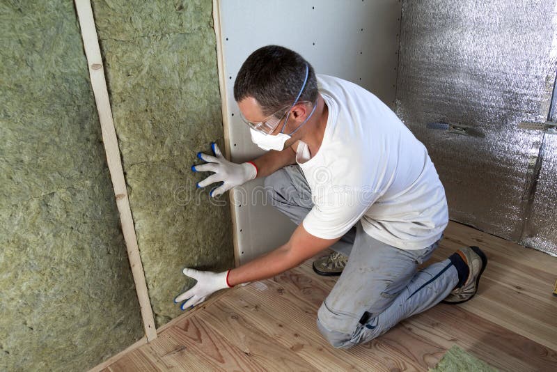 Worker in protective goggles and respirator insulating rock wool insulation in wooden frame for future house walls for cold barrier. Comfortable warm home, economy, construction and renovation concept