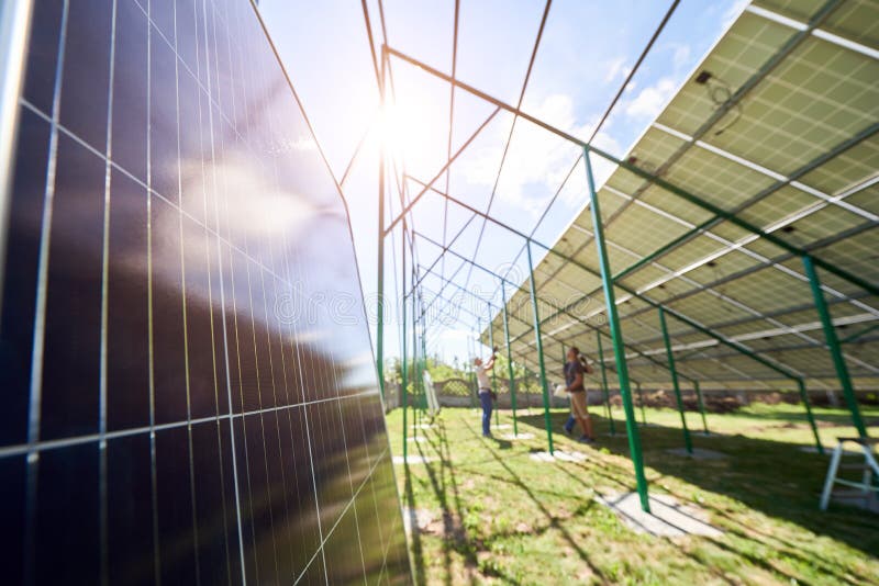 Focus on a solar panel. On blurred background workers mounting innovative solar panels on green metal construction on a sunny day. Providing new technologies for ecology saving, green energy solution