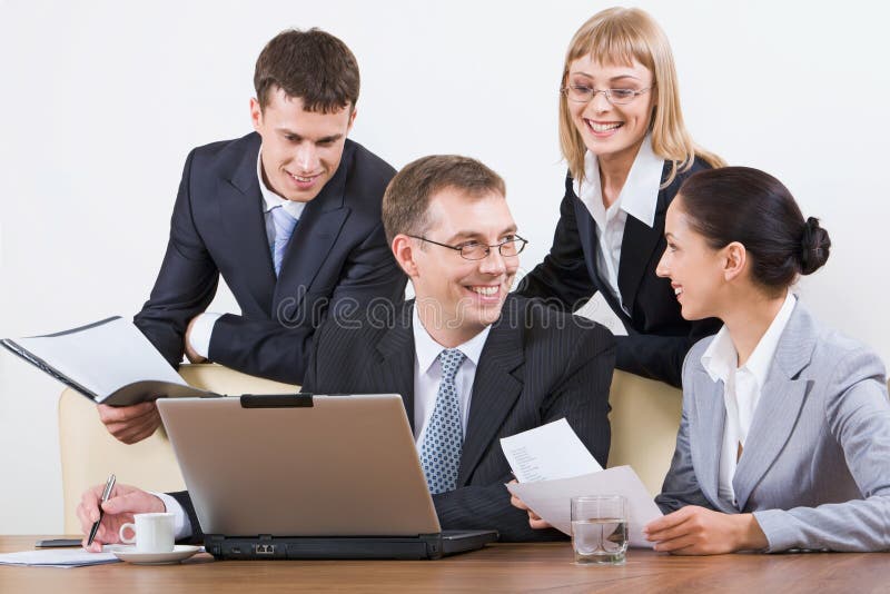 Working in a group. Group of four young businesspeople discussing different questions holding documents gathered together around the table with the laptop Working in a group. Group of four young businesspeople discussing different questions holding documents gathered together around the table with the laptop