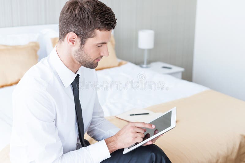 Confident young businessman in shirt and tie working on digital tablet while sitting on the bed in hotel room. Confident young businessman in shirt and tie working on digital tablet while sitting on the bed in hotel room