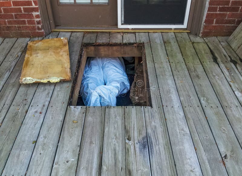 Workman with protective suit crawing under house from crawlspace underneath a wooden deck - only his legs and feet showing