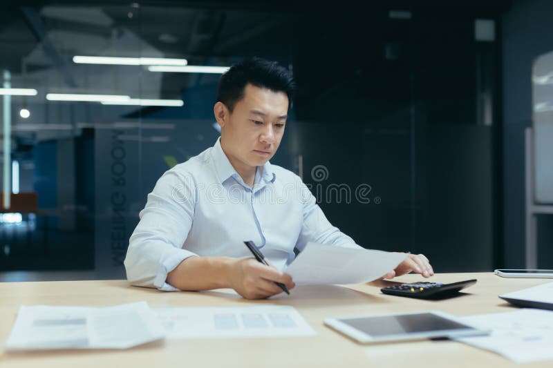 Worried and serious young Asian businessman working in office with documents, busy with paper work, checking accounts on