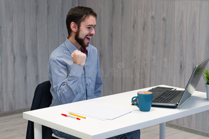 Wow thats a huge win. Young businessman sitting office, looking at laptop display and smiling, clench fists relish some good news