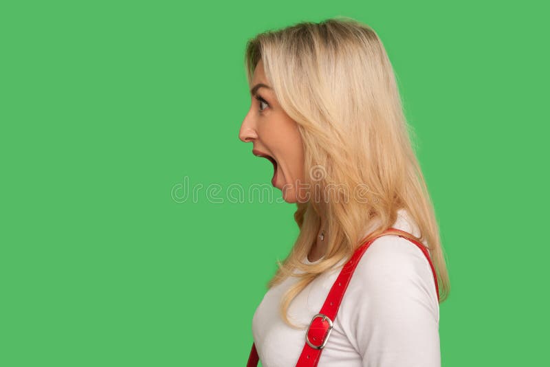 Wow, unbelievable! Side view of astonished adult blond woman standing with wide open mouth and shouting in surprise, shocked expression. indoor studio shot isolated on green background, copy space