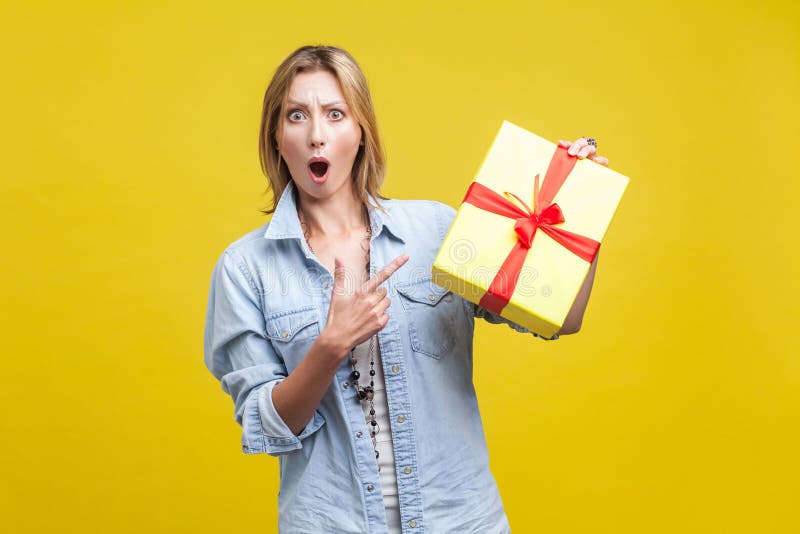 Wow, what a surprise! Portrait of amazed woman pointing at wrapped gift box. studio shot  on yellow