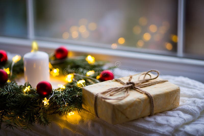 Wrapped gift box near fir wreath decorated with red Christmas balls, white burning candle and coiled with glowing garland with war stock image Wrapped gift box near fir wreath decorated with red Christmas balls, white burning candle and coiled with glowing garland with war stock image