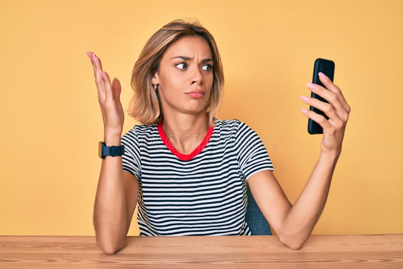 Beautiful caucasian woman doing video call waving to smartphone clueless and confused expression. doubt concept. Beautiful caucasian woman doing video call waving to smartphone clueless and confused expression. doubt concept