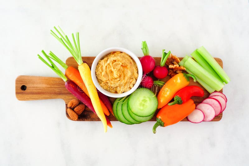 Selection of fresh vegetables and hummus dip on a serving tray. Above view on a white marble background. Selection of fresh vegetables and hummus dip on a serving tray. Above view on a white marble background