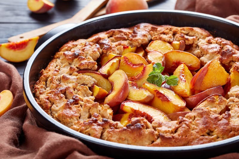 Comfort food dessert: peach crumble in a round black baking dish, close up, with fresh peaches behind the dish. Comfort food dessert: peach crumble in a round black baking dish, close up, with fresh peaches behind the dish