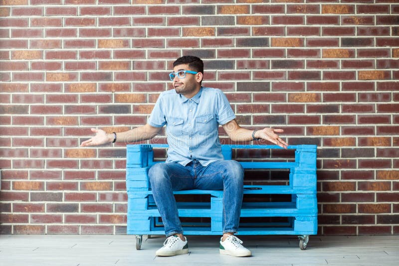 I don`t know. Full length portrait of confused handsome young bearded man in casual style and eyeglasses sitting on blue wooden pallet, looking away and puzzled. indoor studio shot on brown brick wall. I don`t know. Full length portrait of confused handsome young bearded man in casual style and eyeglasses sitting on blue wooden pallet, looking away and puzzled. indoor studio shot on brown brick wall