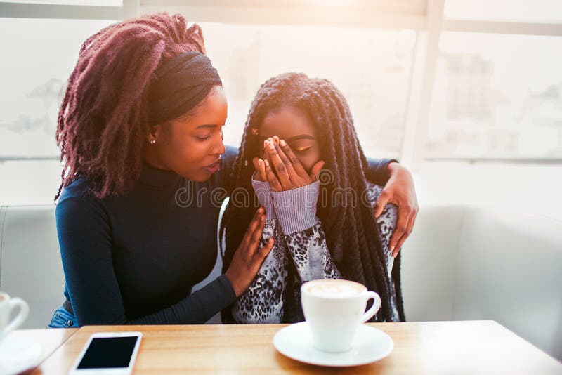 Wzburzonego youn afrykańska kobieta siedzi i płacze Zakrywa twarz z rękami Jej przyjaciel wygody młoda kobieta Siedzą w kawiarni