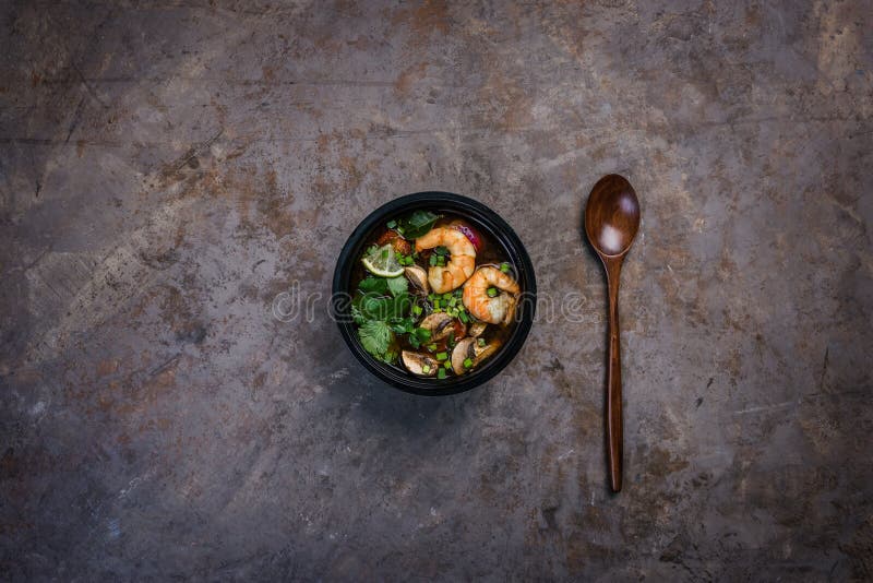 Würzige thailändische Suppe Tom Yam mit Meeresfrüchten in einer schwarzen Schüssel auf einer Weinlese färbte Hintergrund mit eine stockbild Würzige thailändische Suppe Tom Yam mit Meeresfrüchten in einer schwarzen Schüssel auf einer Weinlese färbte Hintergrund mit eine stockbild