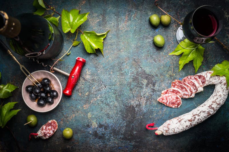 Italian still life with salami, red wine, olives and grape leaves on dark rustic background, top view, place for text. Italian food background. Italian still life with salami, red wine, olives and grape leaves on dark rustic background, top view, place for text. Italian food background