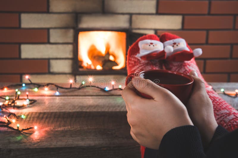Xícara de café vermelha na mão fêmea pela chaminé A fêmea relaxa pelo warmfire em peúgas do vermelho do Natal Feriado do Natal imagens de stock Xícara de café vermelha na mão fêmea pela chaminé A fêmea relaxa pelo warmfire em peúgas do vermelho do Natal Feriado do Natal imagens de stock