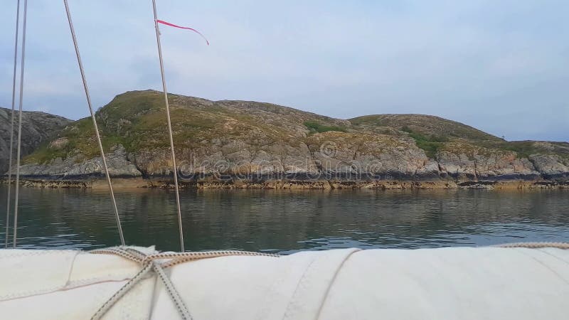 Yacht sailing along stony shore in calm waters. Red ribbon tied to a cable waives in wind.