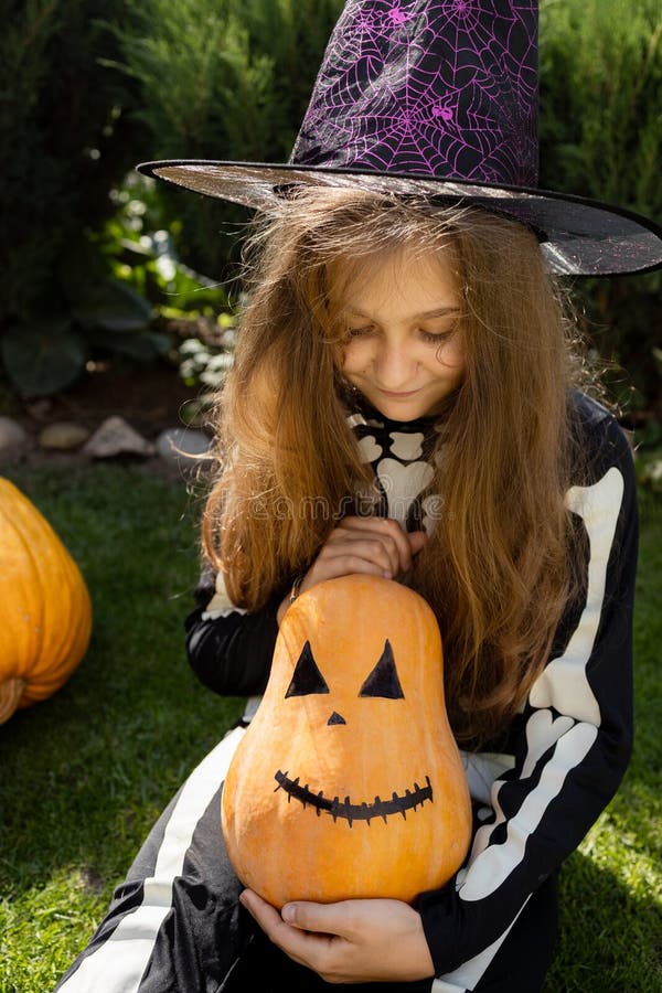 10-year-old girl in a skeleton costume and a witch hat holds in her hands a Halloween pumpkin with a drawn grin