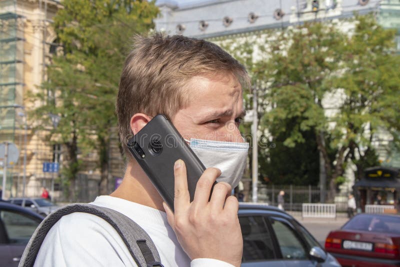 A 20-25-year-old guy in a medical mask is talking on a mobile phone against the background of a European city. Concept: studying i royalty free stock image A 20-25-year-old guy in a medical mask is talking on a mobile phone against the background of a European city. Concept: studying i royalty free stock image