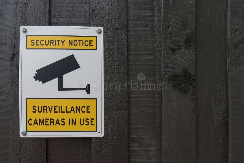 A yellow, black and white Security Notice, Surveillance Cameras In Use warning sign on a painted wooden fence
