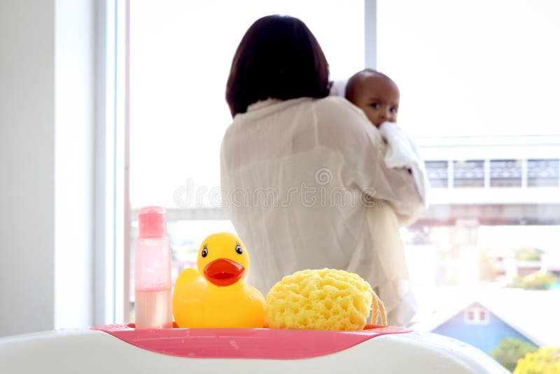 Yellow duck toy, shampoo soap and sponge baby toiletries on bathtub in bathroom with blurry background of mother holding infant