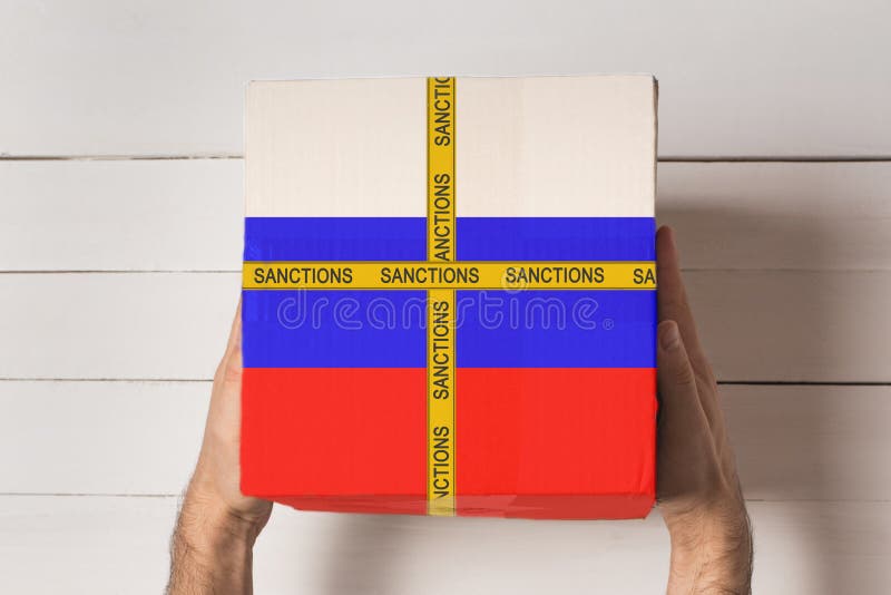 Yellow tape with the inscription Sanctions on the box with the flag of Russia in male hands. Top view White table in the stock images Yellow tape with the inscription Sanctions on the box with the flag of Russia in male hands. Top view White table in the stock images