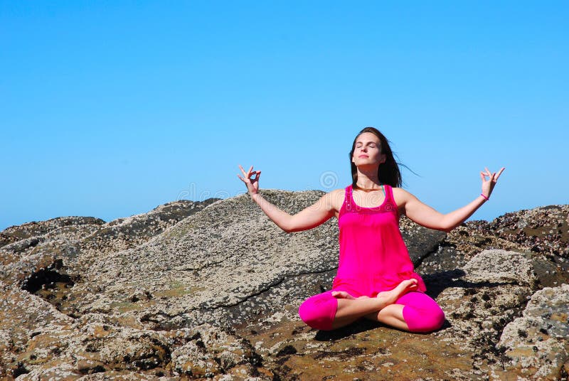 A beautiful young woman in pink clothes with relaxed facial expression sitting on the rocks on a beach, doing yoga in nature outdoors. A beautiful young woman in pink clothes with relaxed facial expression sitting on the rocks on a beach, doing yoga in nature outdoors