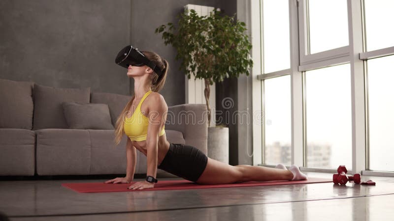 Yogi kobieta uprawia poranne ćwiczenia sportowe w domu rozciągające się na czerwonej matie, nosząc maskę VR Elastyczna dziewczyna