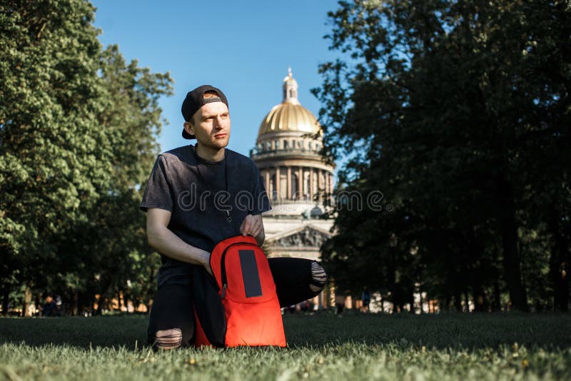 Youg-Manntourist mit dem Rucksack, der auf Picknick auf Gras nahe Heilig-Isaac-` s Kathedrale in St Petersburg in sonnigem sitzt stockfotografie