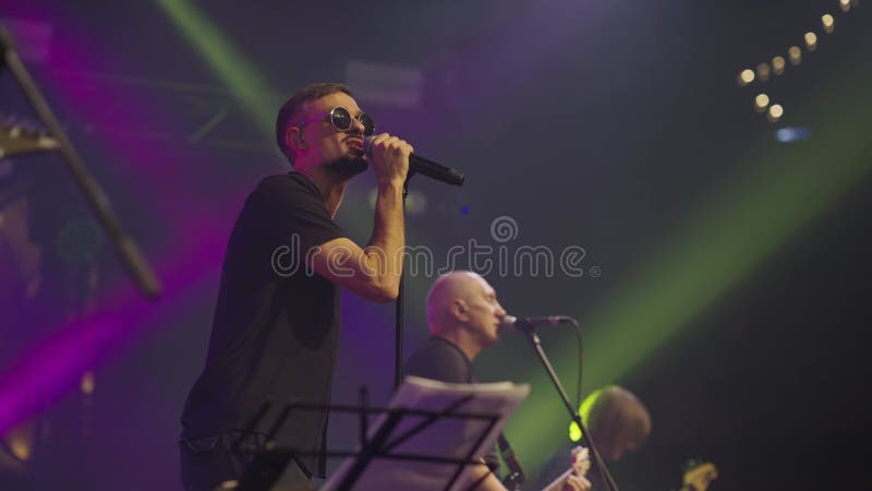 Young active artist sings into microphone and gestures with his hands while performing on stage with group stock video footage