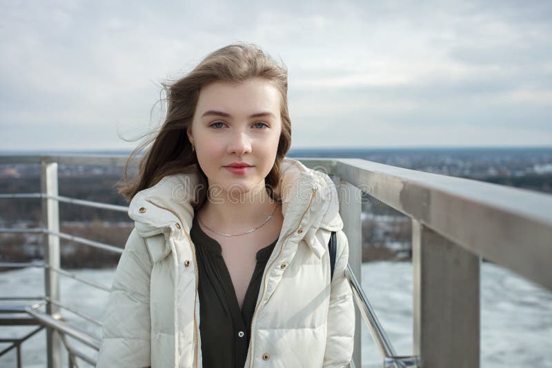 Young adorable smiling blonde teen girl having fun on the observation deck with a view of cloudy spring sky, frozen river, sunny w