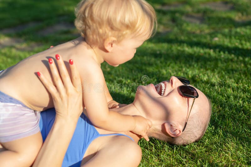 Young adult beautiful bald caucasian mom enjoy lying with little blond toddler boy son on green grass lawn at city park at summer royalty free stock images Young adult beautiful bald caucasian mom enjoy lying with little blond toddler boy son on green grass lawn at city park at summer royalty free stock images
