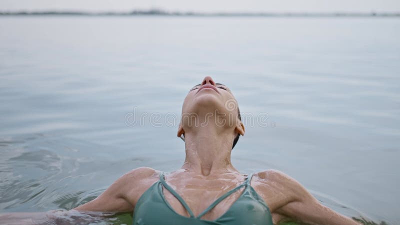 A young adult woman relaxes lying on the water surface in lake, eyes closed contentedly as she enjoys a dip in the water