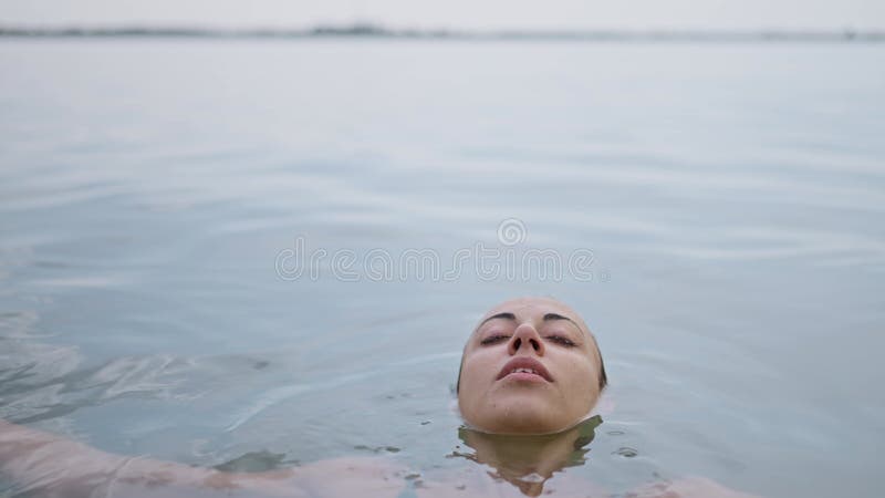 A young adult woman relaxes lying on the water surface in lake, eyes closed contentedly as she enjoys a dip in the water