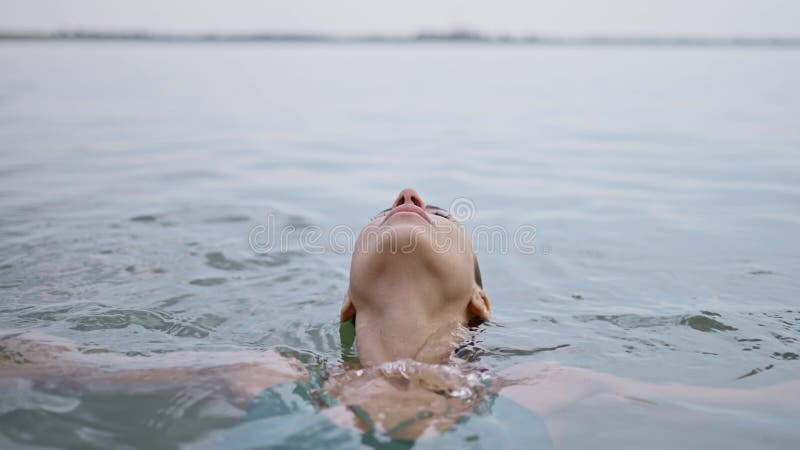 A young adult woman smiles and relaxes lying on the water surface in lake, eyes closed contentedly as she enjoys a dip