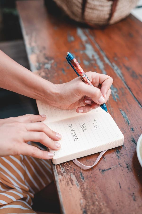 Young adult woman writing in a journal &x22;you are divine&x22; on a wooden table