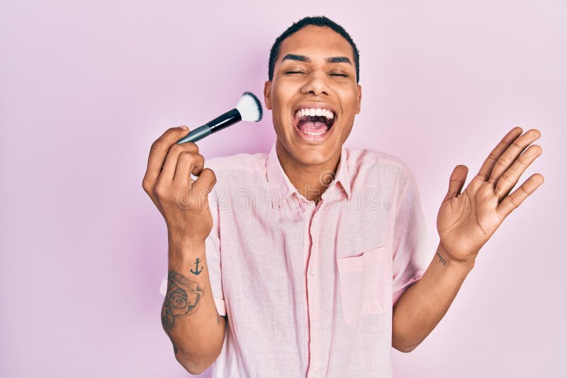 Young african american guy holding makeup brush celebrating achievement with happy smile and winner expression with raised hand royalty free stock images