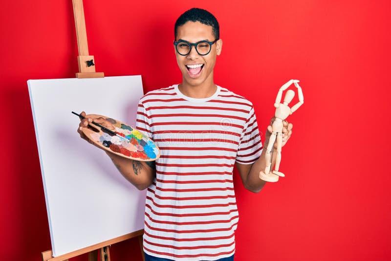 Young african american guy holding painter palette and art manikin winking looking at the camera with sexy expression, cheerful stock photo