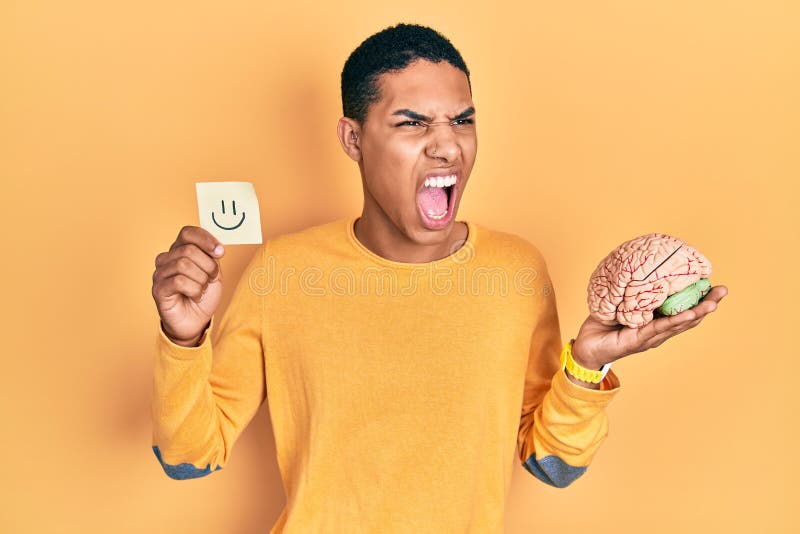Young african american guy holding smile reminder and brain angry and mad screaming frustrated and furious, shouting with anger royalty free stock photos