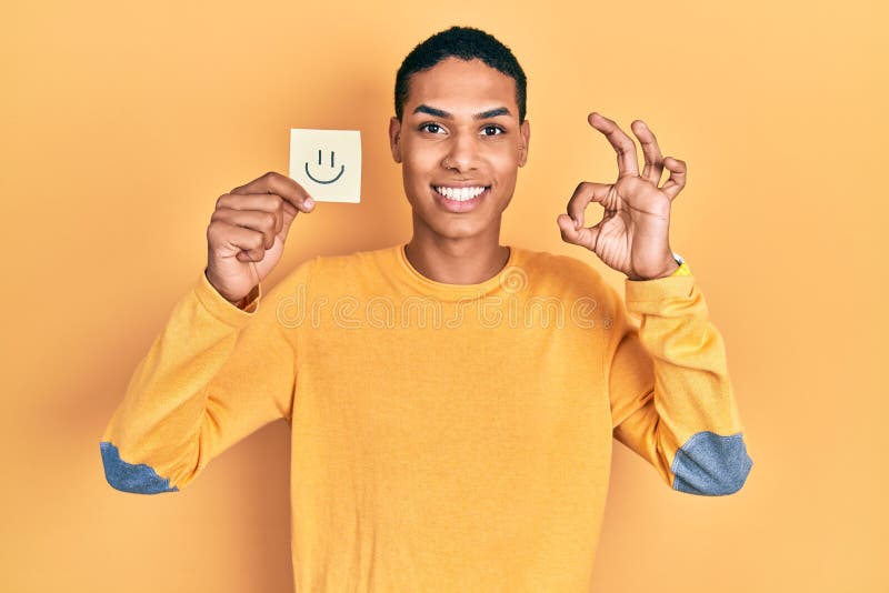 Young african american guy holding smile reminder doing ok sign with fingers, smiling friendly gesturing excellent symbol royalty free stock photography