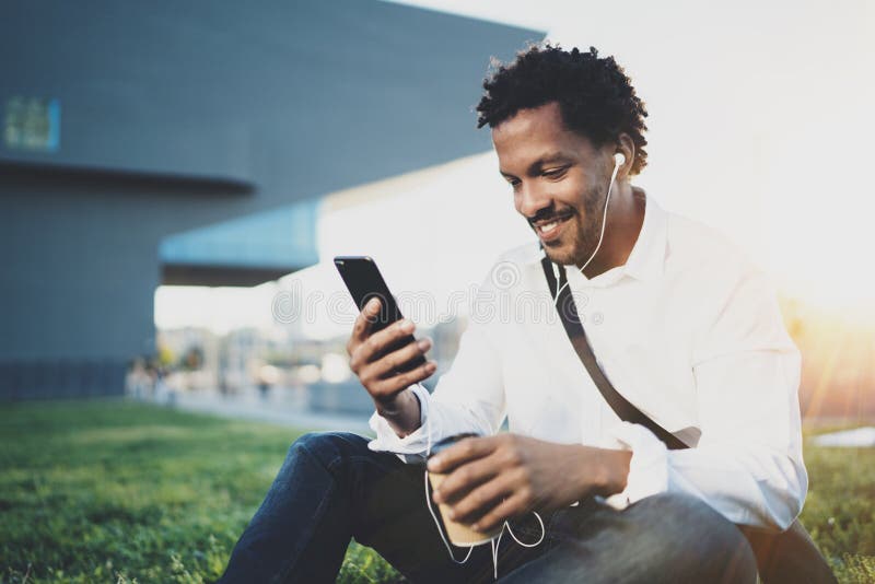 Young African American man in headphone sitting at sunny city park and enjoying to listen to music on his smartphone stock images Young African American man in headphone sitting at sunny city park and enjoying to listen to music on his smartphone stock images