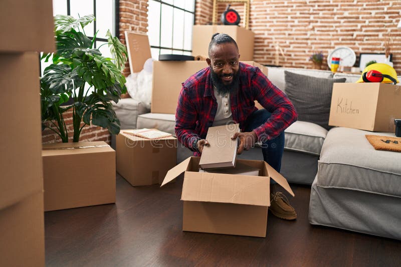 Young african american man unpacking books cardboard box at new home