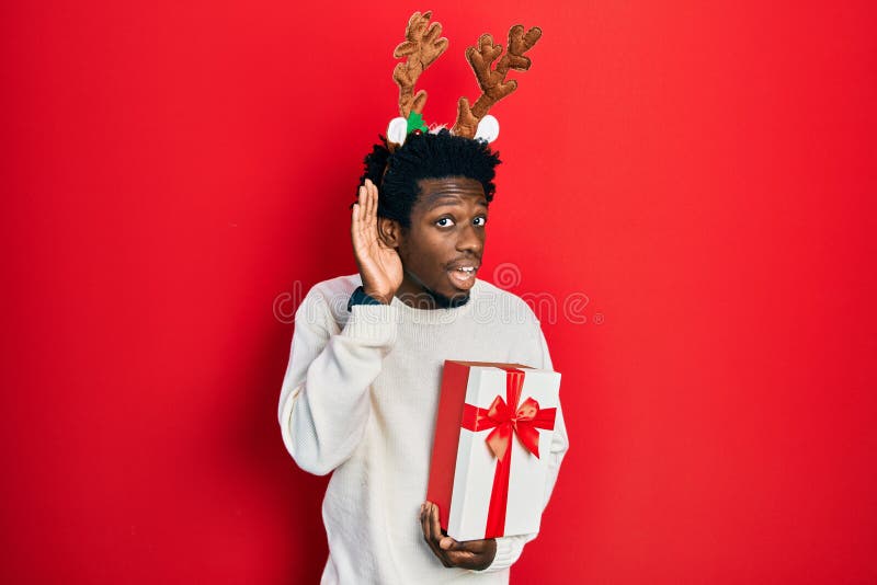 Young african american man wearing deer christmas hat holding gift smiling with hand over ear listening an hearing to rumor or