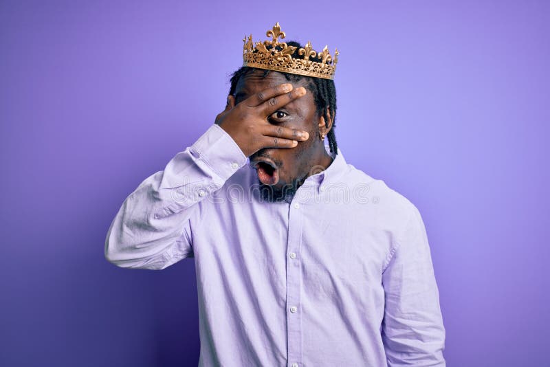 Young african american man wearing golden crown of king over isolated purple background peeking in shock covering face and eyes stock photography