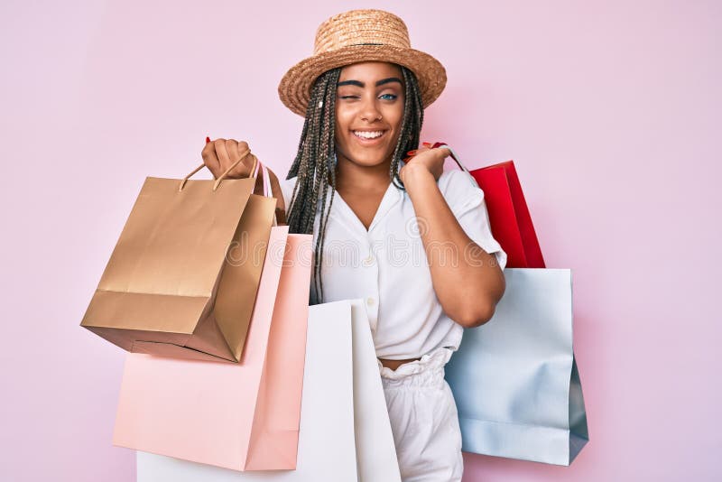 Young african american woman with braids holding shopping bags winking looking at the camera with sexy expression, cheerful and