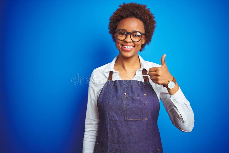 Young african american woman shop owner wearing business apron over blue background doing happy thumbs up gesture with hand