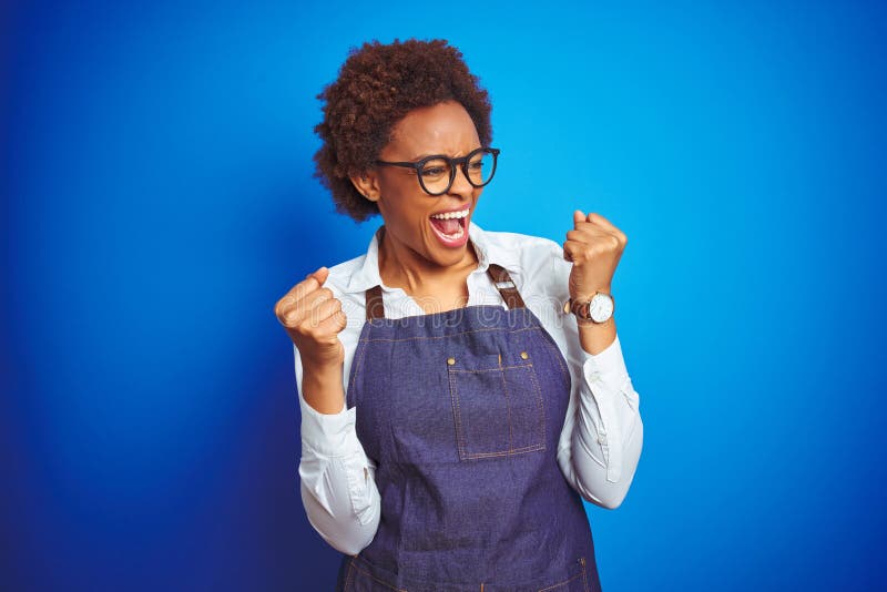 Young african american woman shop owner wearing business apron over blue background very happy and excited doing winner gesture royalty free stock photography