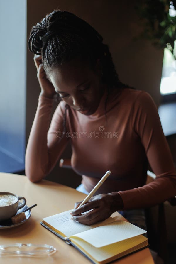 Young African-American woman sitting at beige table with cup of coffee, wooden tray in cafe, writing text in notebook. royalty free stock images