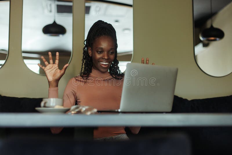 Young African-American woman sitting at table in cafe near laptop, raising hands up, expressing success, win, greeting. royalty free stock photography