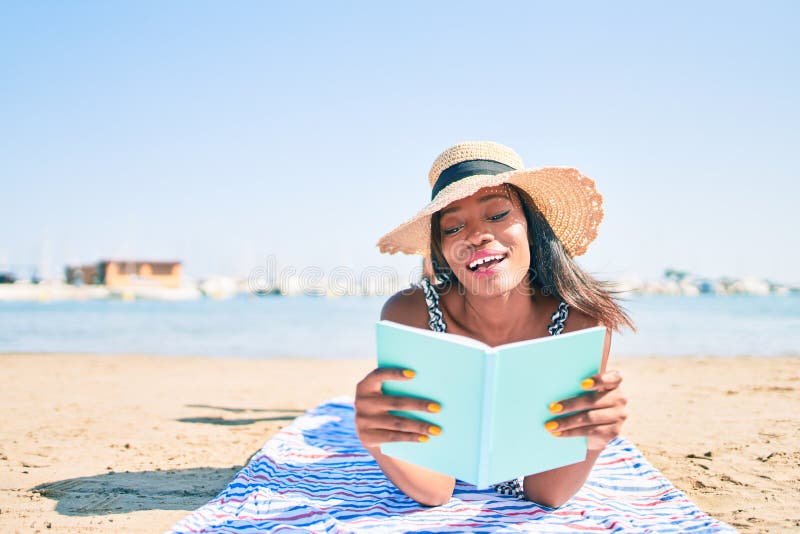 Young african american woman on vacation laying on the towel reading book at the beach stock photo