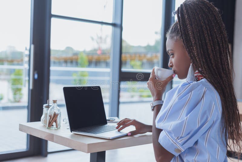 Young african american woman working with laptop and drinking coffee at cafe